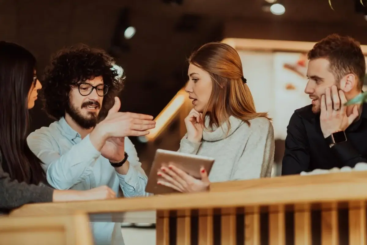 Four coworkers sit around a wooden railing in a cafe, one man gestures as he explains while a woman holds a tablet and listens, others observe.