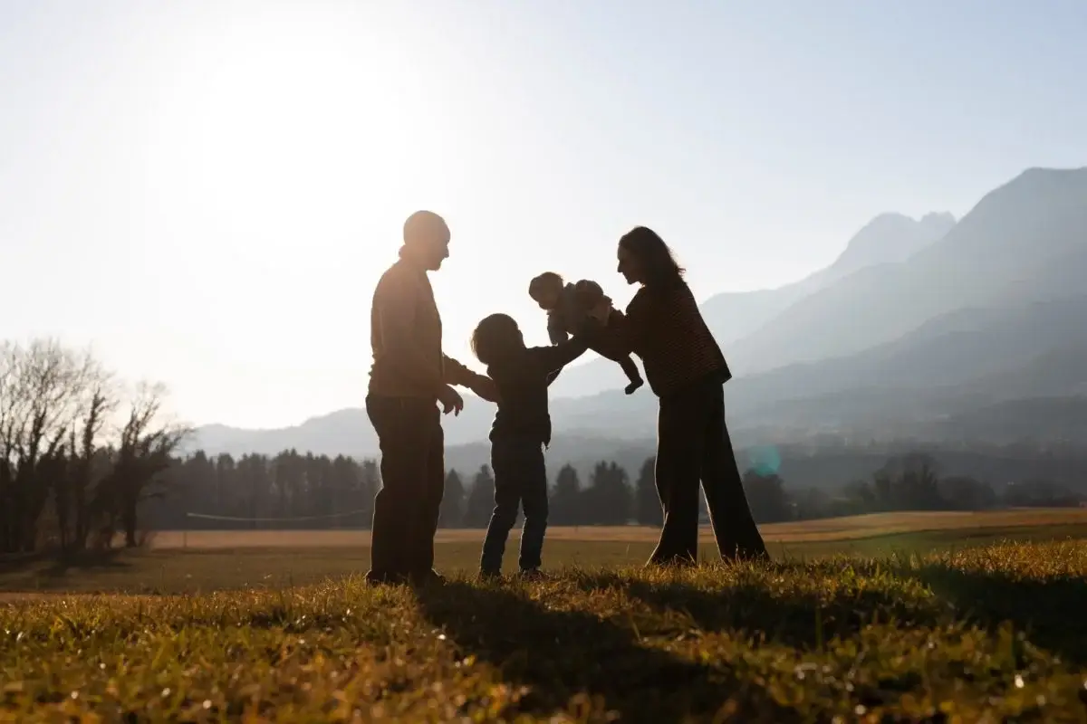 Silhouetted family of four standing in a sunlit field, lifting a baby between them with mountains in the background.