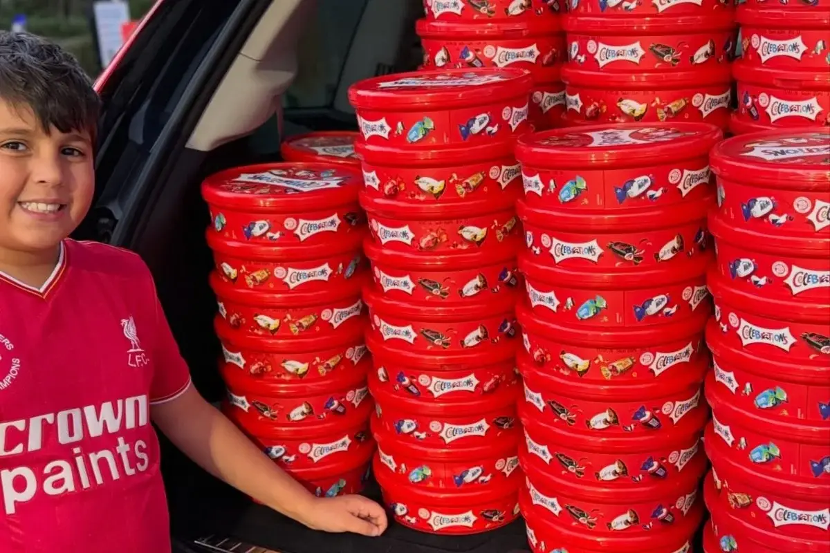 Smiling boy in a pink Crown Paints shirt stands beside tall stacks of red paint buckets labeled 'Celebations.'