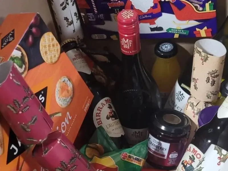 A cluttered shelf of festive items: wine bottle, jam jar, crackers, and assorted gift boxes.