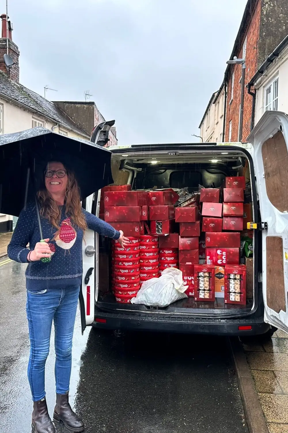 Woman smiling under a large black umbrella beside an open van loaded with red boxes and bags in the rain on a street.”,