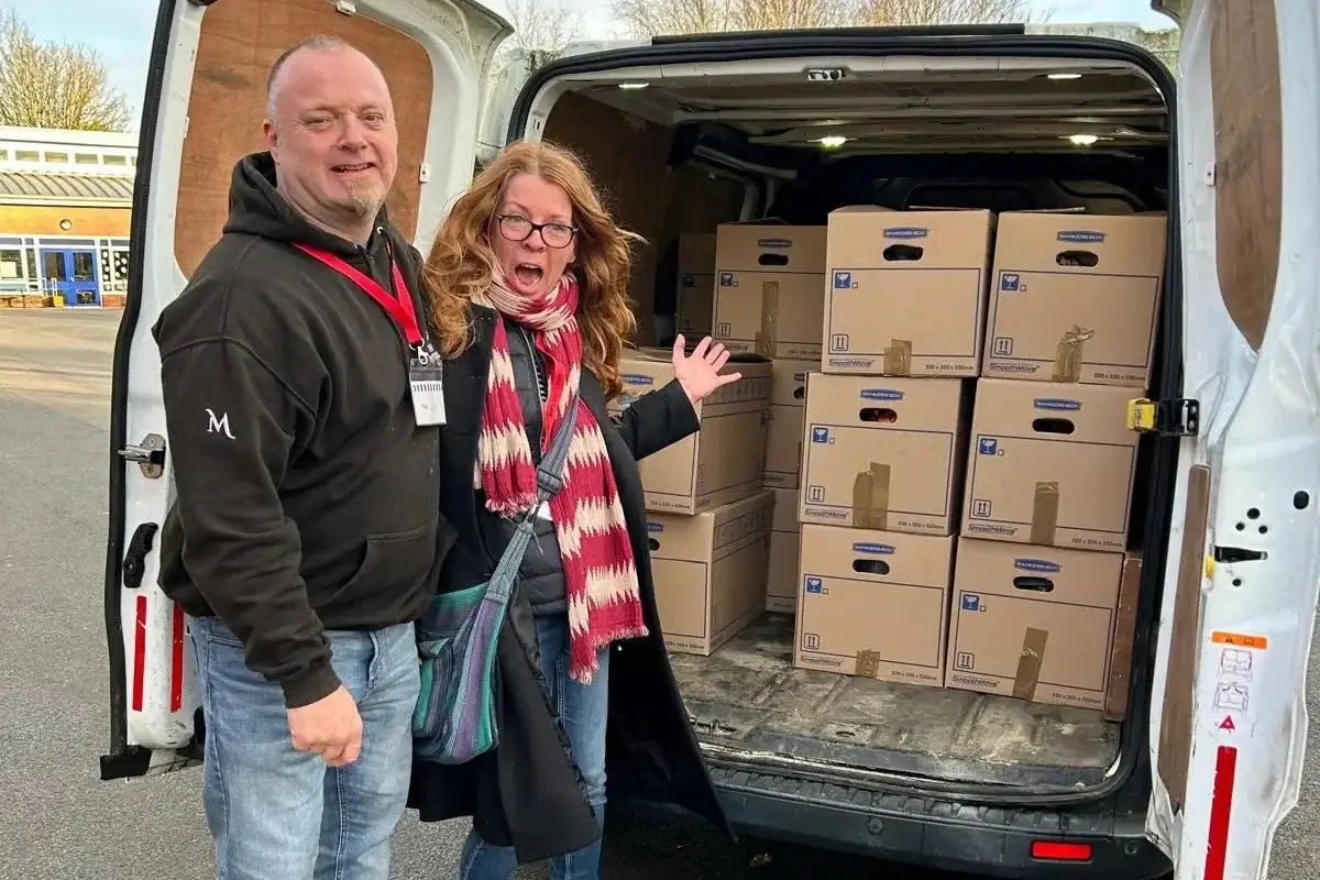 Two people stand beside an open van loaded with stacked cardboard boxes inside and out in a parking lot, smiling at the camera.