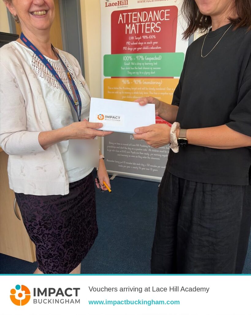 Two women exchange a white voucher envelope during a visit to Lace Hill Academy, with the Impact Buckingham logo visible on the envelope and a promotional poster in the background.