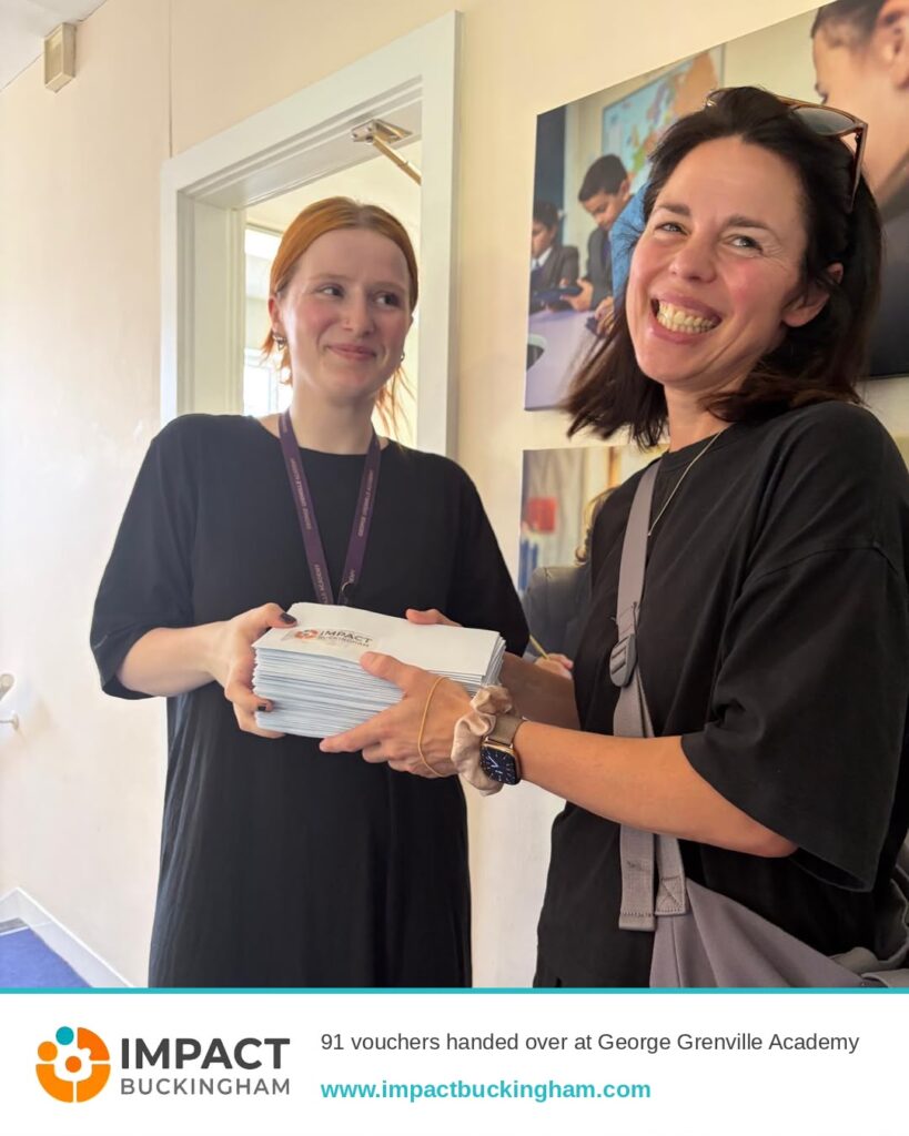 Two women in black shirts exchange a stack of vouchers, both smiling, during a charity event with Impact Buckingham branding visible at the bottom.